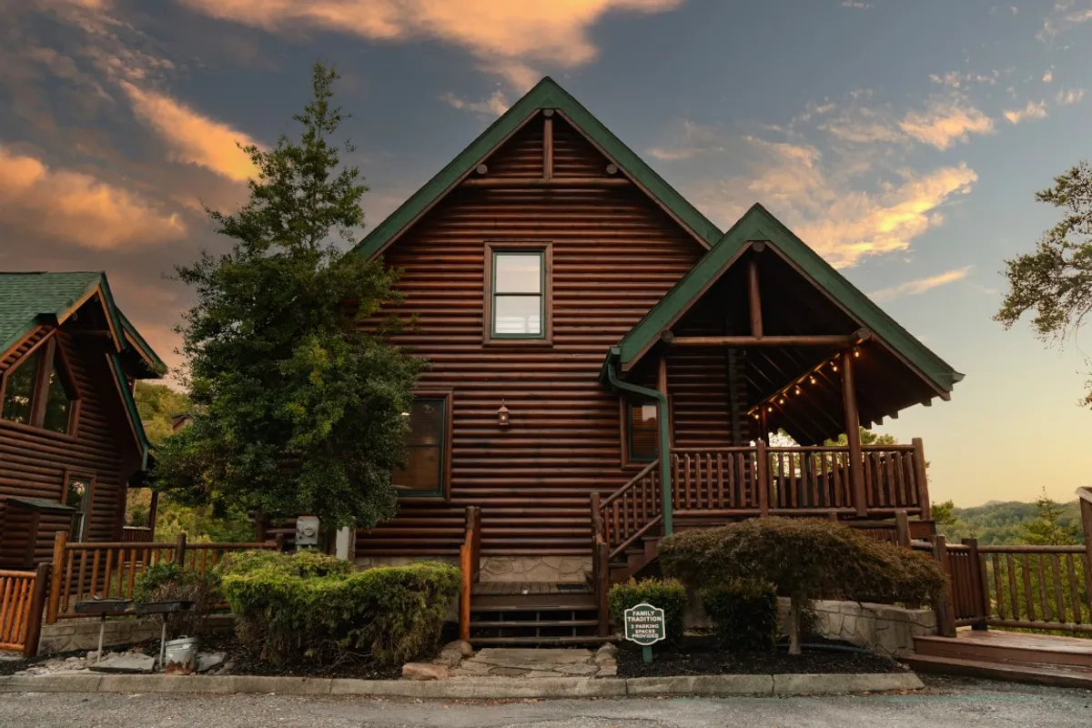The image showcases a classic log cabin with a green roof nestled among lush foliage. An expansive wooden deck wraps around the cabin, inviting guests to relax and take in sweeping mountain vistas. The warm, natural tones of the wood and the surrounding trees create a cozy, welcoming ambiance, perfect for family gatherings and outdoor relaxation.
