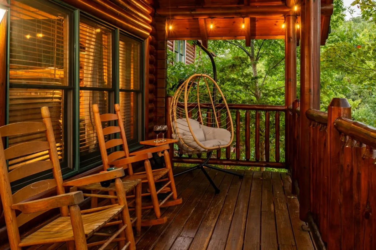 A cozy outdoor porch setup featuring a hanging chair, wooden rocking chairs, an outdoor table, and glowing string lights. The porch is surrounded by trees, creating a secluded and inviting spot to relax, enjoy a drink, or read a book.