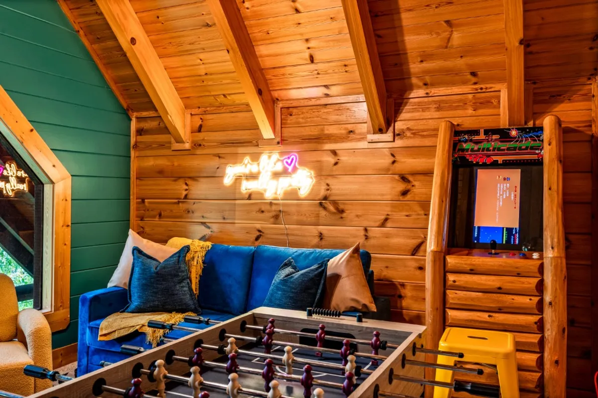 A fun-packed game room with a blue couch, foosball table, arcade game machine, and a neon sign, presenting a relaxed area for both play and chilling with friends. The arrangement fosters both active gaming and laid-back socializing.