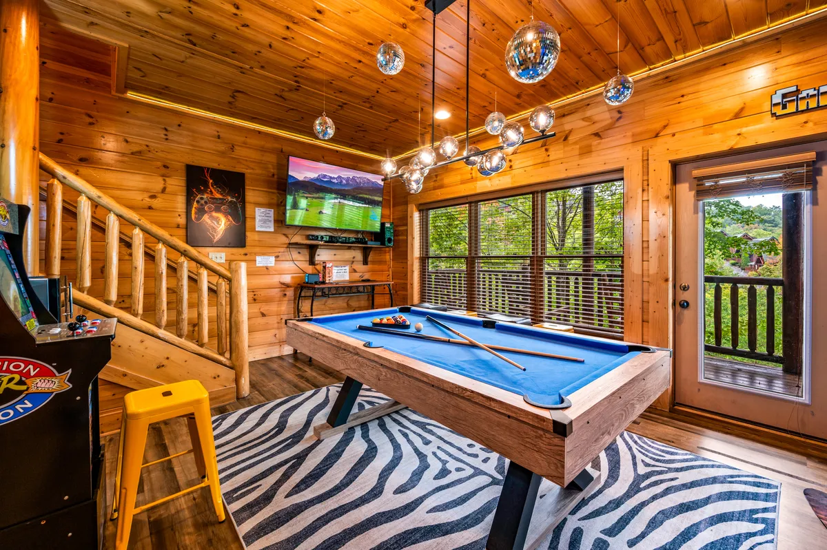 A vibrant game room featuring a pool table in the center atop a striking zebra rug, arcade game machine in the corner, disco balls hanging from the ceiling, and a large television mounted on the wall. The overall decor is playful and energetic, perfect for group activities and celebrations.