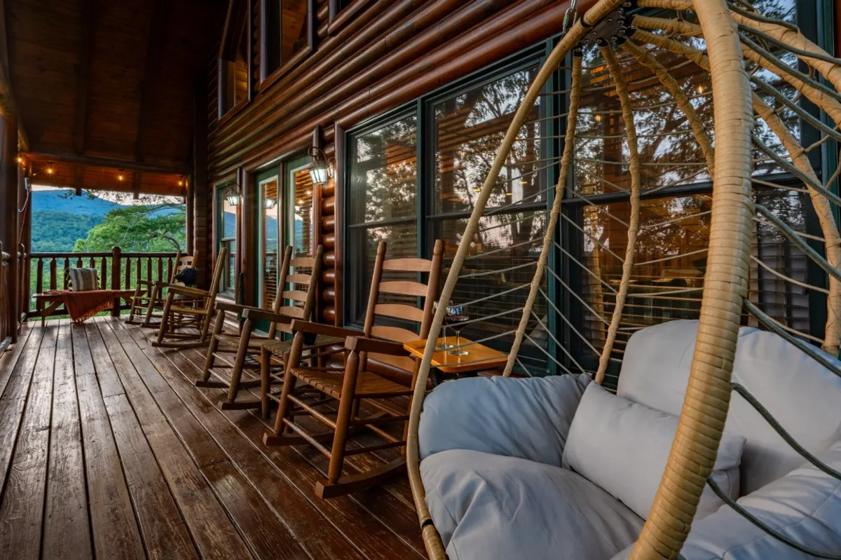 A spacious wooden deck extends outward, furnished with a hanging chair and several classic wooden rocking chairs. The deck overlooks a breathtaking mountain view, with lush treetops stretching beneath a vast sky. The setting radiates calm, offering a harmonious blend of comfort and nature.