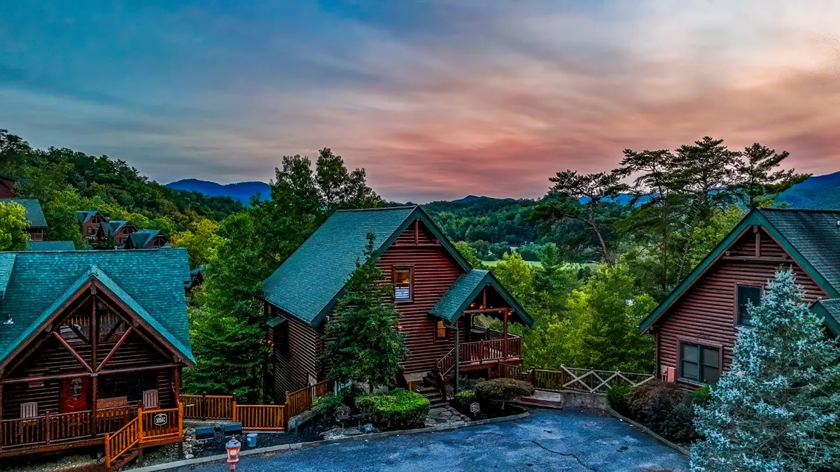 A scenic shot of multiple log cabins set among trees, with a mountain backdrop and the sky painted in sunset hues. The open, expansive setting suggests both community and adventure, highlighting the Smoky Mountains' natural beauty and the proximity to outdoor activities.