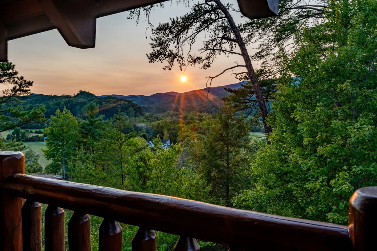 A stunning view of the sun setting over the Smoky Mountains, with lush greenery in the foreground and a rustic wooden railing framing the scene. The fading daylight bathes the landscape in a tranquil glow, inviting guests to pause and appreciate the natural beauty.
