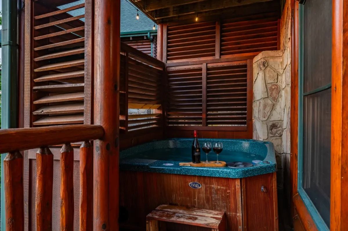 A private hot tub is set on the deck, framed by rustic wooden railings. A wine bottle and glasses rest on the edge, inviting guests to unwind with a drink. The backdrop features the soft glow of sunset over distant mountains, enhancing the sense of privacy and indulgence.