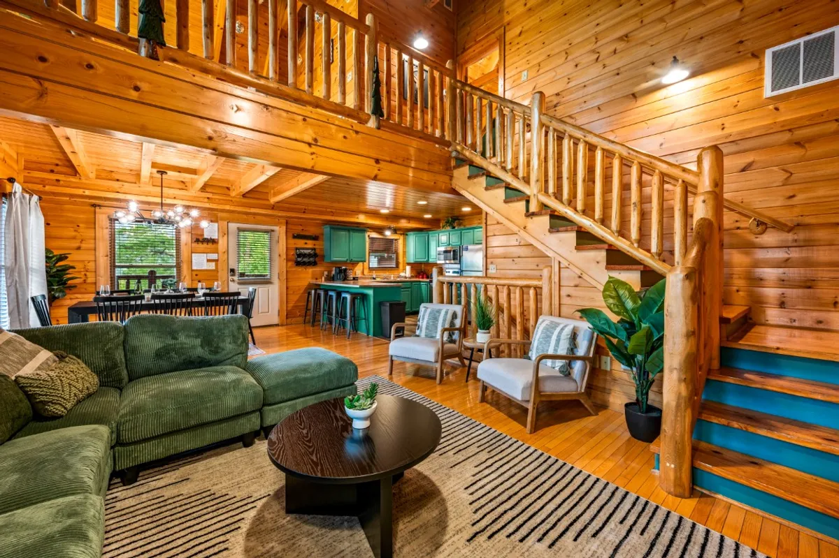 This photo shows the heart of the cabin, with a green sectional sofa in the living area, a sturdy wooden dining table for group meals, and the adjacent kitchen equipped with bar stools and modern appliances. Decorative plants and a wooden staircase add a cozy touch, while the open floor plan emphasizes togetherness.
