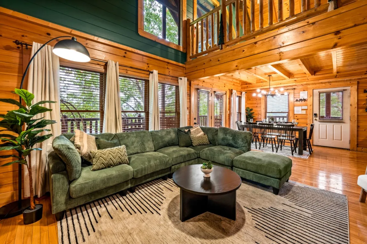 A wide view of the open-concept living area, featuring a large green sectional sofa, a round black coffee table, and the dining area in the background. Sunlight pours in through expansive windows, and exposed wooden beams give a warm, rustic feel to the space. The seamless transition between the living and dining areas is clearly visible, emphasizing the spaciousness and sociability of the room.
