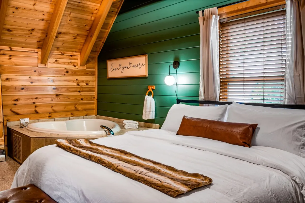 A cozy bedroom featuring a plush bed with luxurious linens, a jacuzzi tucked beside wooden accents, and a decorative sign that adds a personal touch. Warm, intimate lighting enhances the inviting atmosphere, making the room ideal for relaxation and unwinding after a day of adventure.