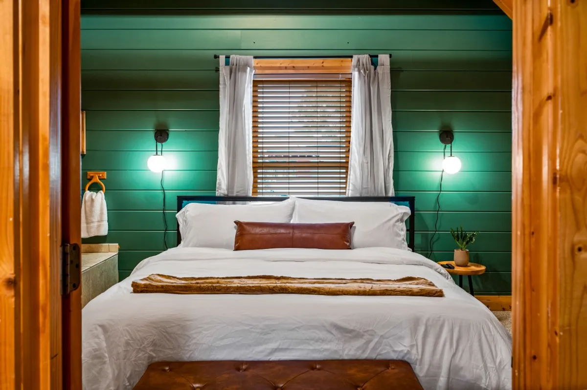 A tranquil guest bedroom with crisp white bedding, a green wooden accent wall, wooden nightstand with a potted plant, and a wooden bench at the foot of the bed. Natural light streams in, illuminating the calm, nature-inspired decor and inviting a sense of peace.