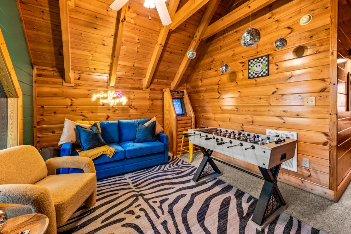 A vibrant game room inside the cabin features a blue sofa, foosball table, disco balls, an arcade game, and a zebra rug. The playful decor and layout encourage friendly competition, laughter, and bonding, aligning with the section’s mention of two game rooms and group social activities.